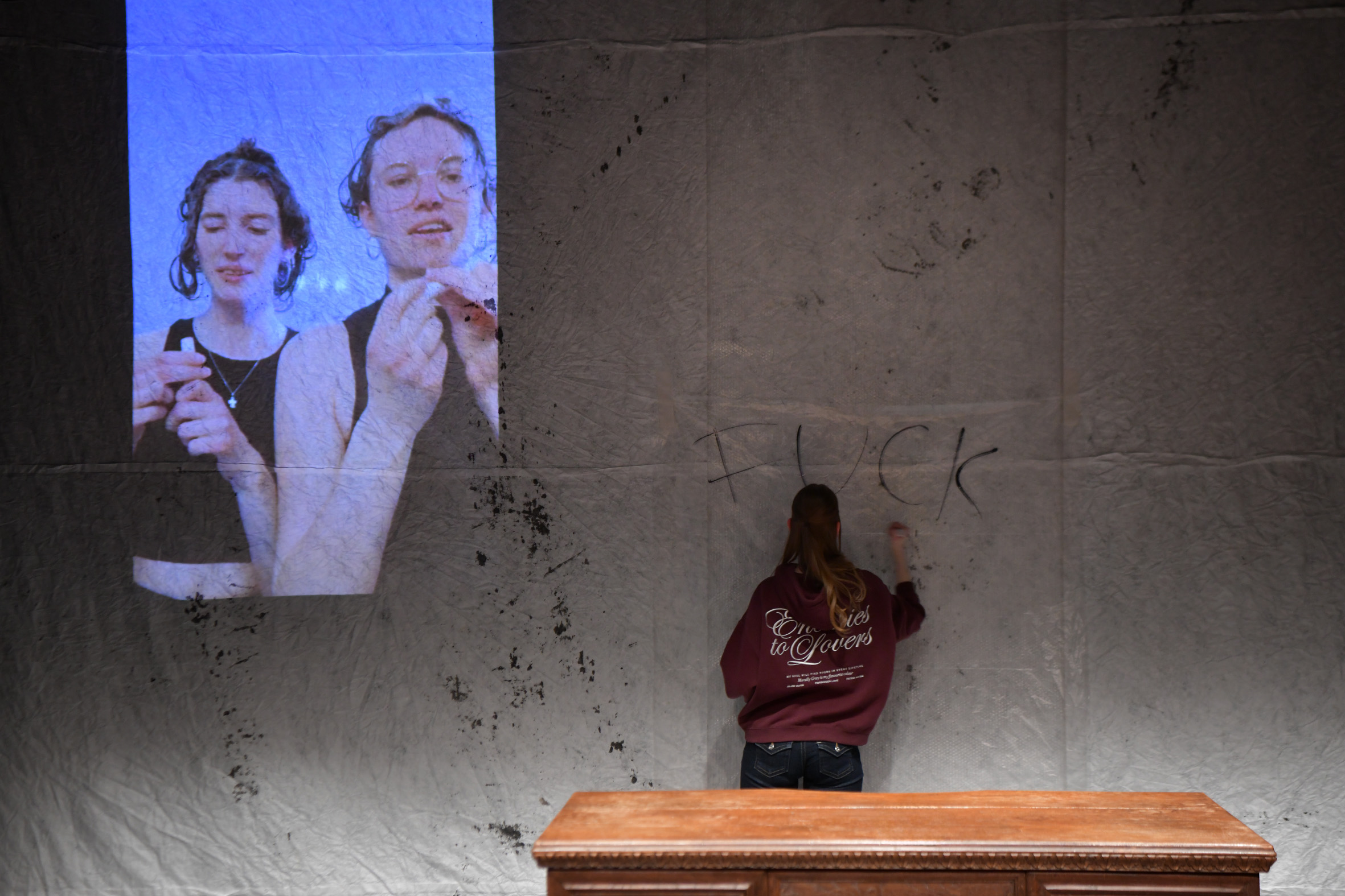 Mittig steht eine Person, die mit Edding "Fuck you" an die fleckige helle Wand schreibt. Links als Videoprojektion: Zwei Darstellerinnen verwenden Makeup und haben Shampoo im Haar. 